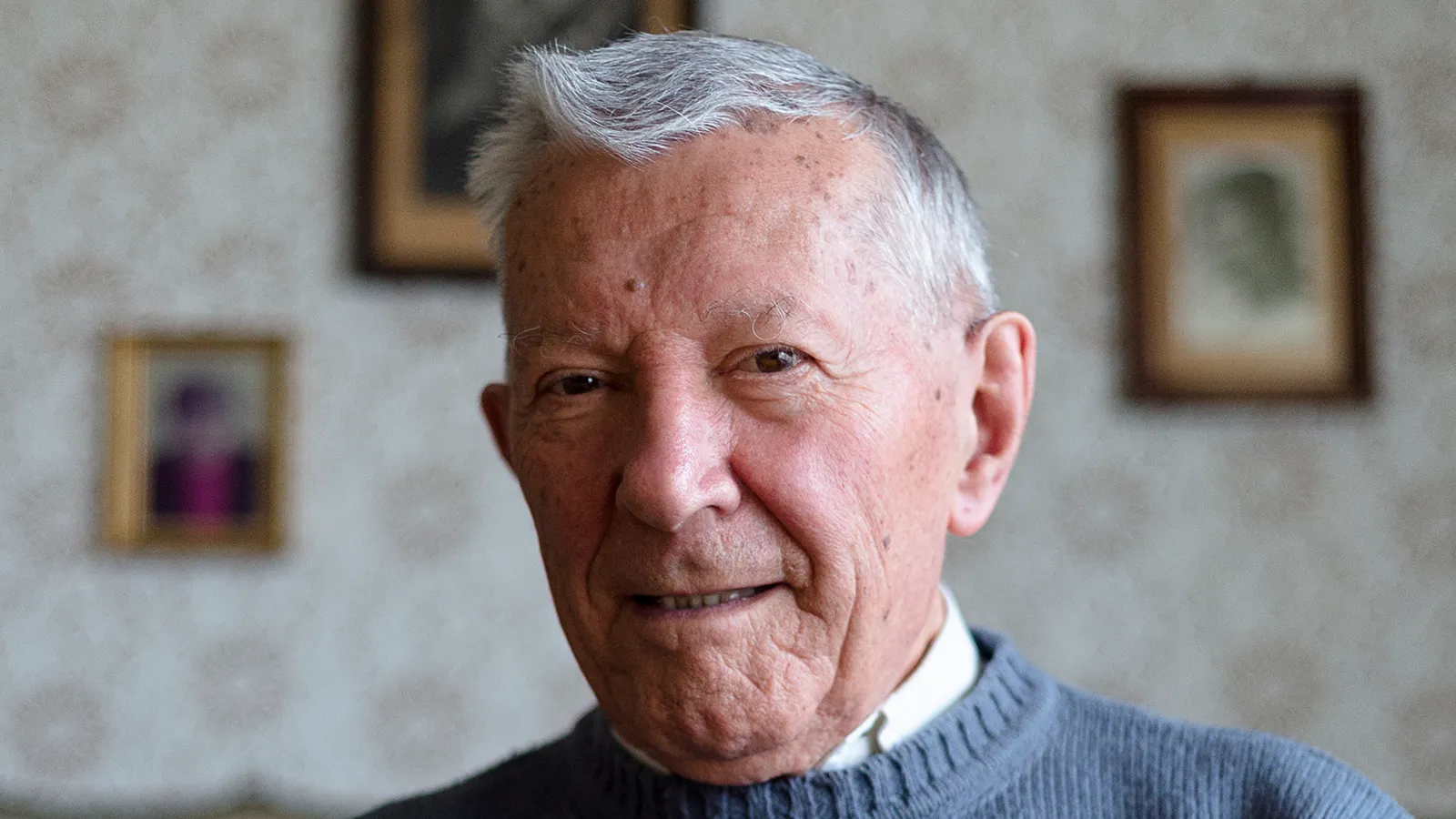 Älterer Mann sitzt im Wohnzimmer - hinter ihm sind alte Bilder an der Wand