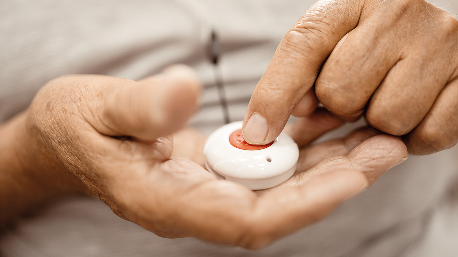 Zwei alte Hände, in einer Hand liegt der Hausnotruf-Knopf, mit dem Zeigefinger der anderen Hand wird auf den Knopf gedrückt