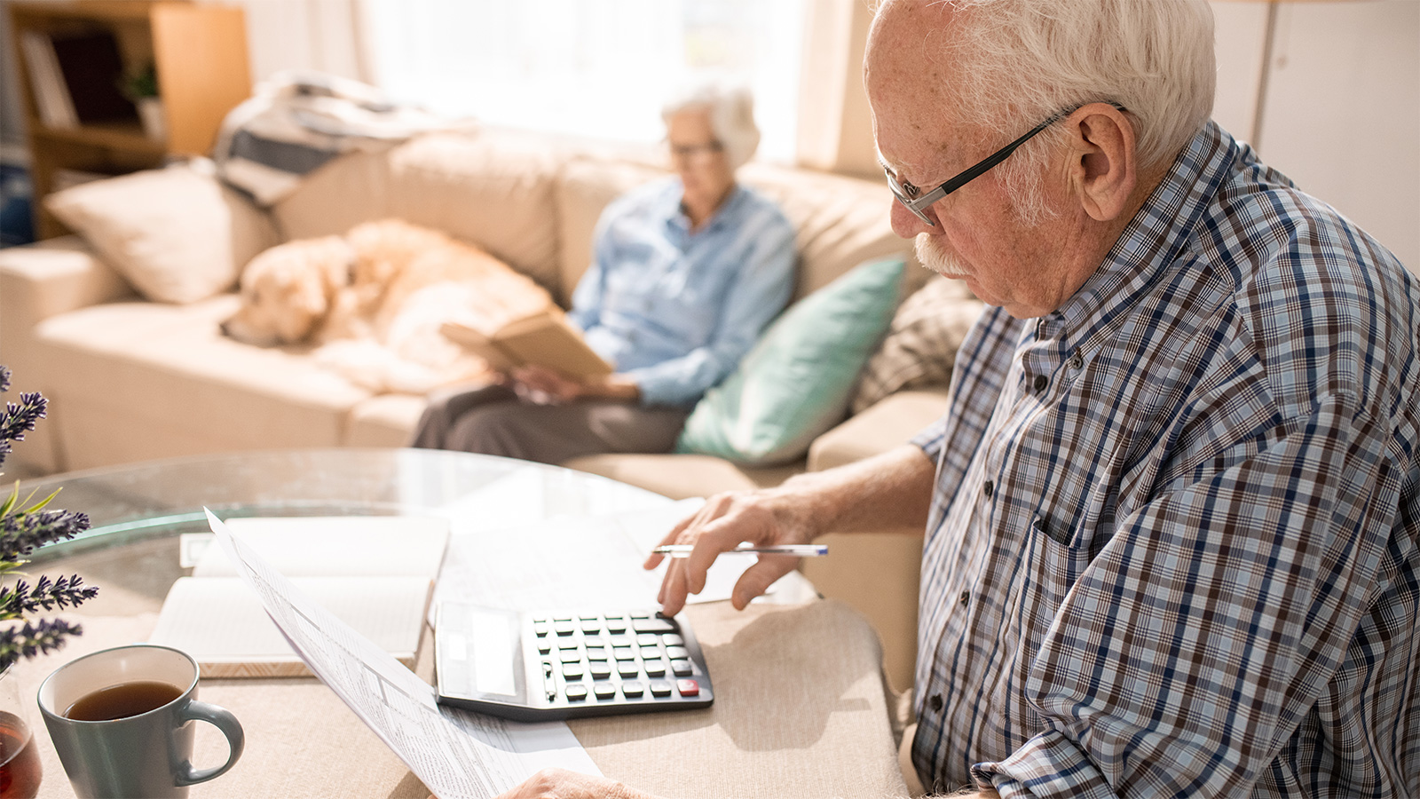 Senior sitzt am Tisch und tippt auf einen Taschenrechner. In der anderen Hand hält er Formulare. Im Hintergrund sitzt eine ältere Dame auf dem Sofa und liest.