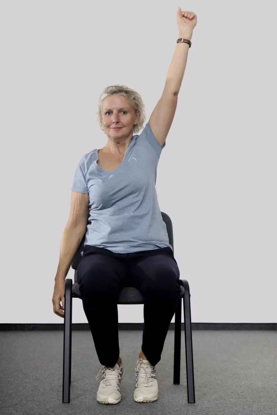 Frau sitzt auf einem Stuhl und reckt den linken Arm in die Höhe