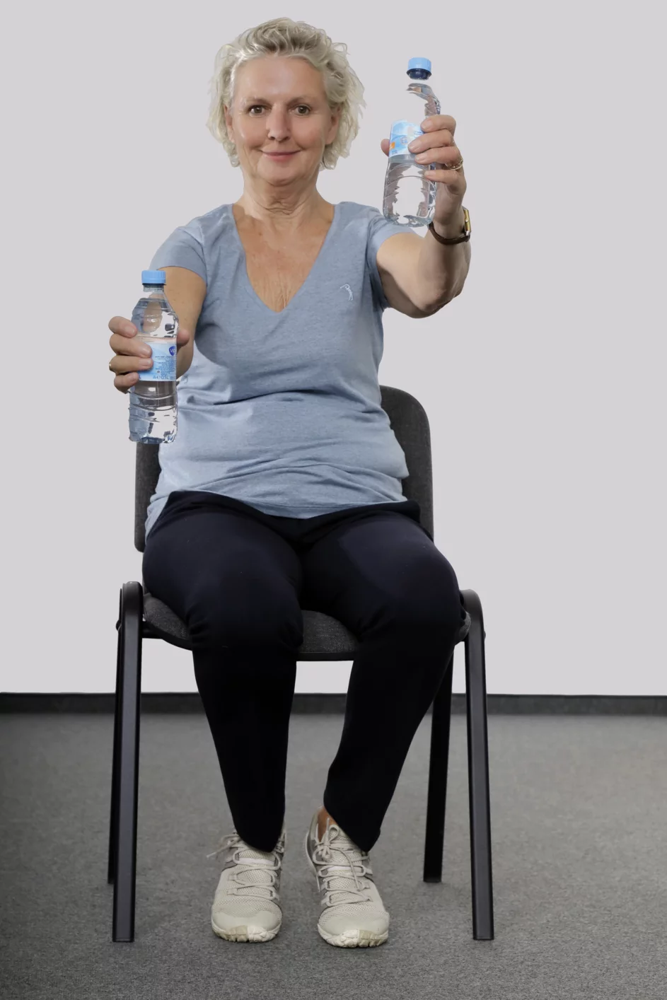 Frau sitzt auf einem Stuhl, hat in jeder Hand eine Wasserflasche und streckt abwechselnd die Arme nach vorne