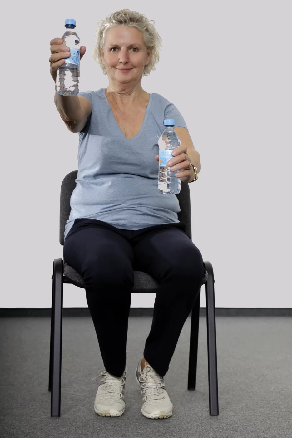Frau sitzt auf einem Stuhl, hat in jeder Hand eine Wasserflasche und streckt abwechselnd den rechten und den linken Arm nach vorne aus