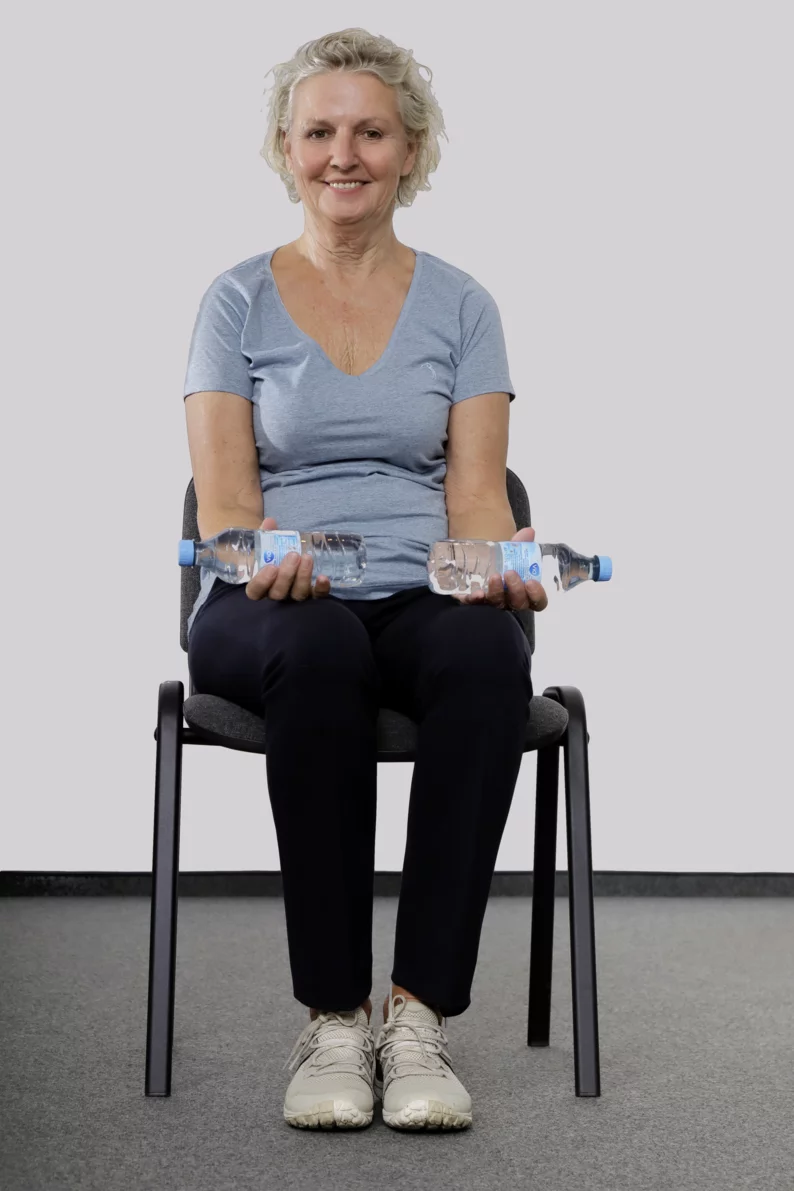 Frau sitzt auf einem Stuhl und hat in jeder Hand eine Wasserflasche. Die Hände liegen auf den Oberschenkeln.