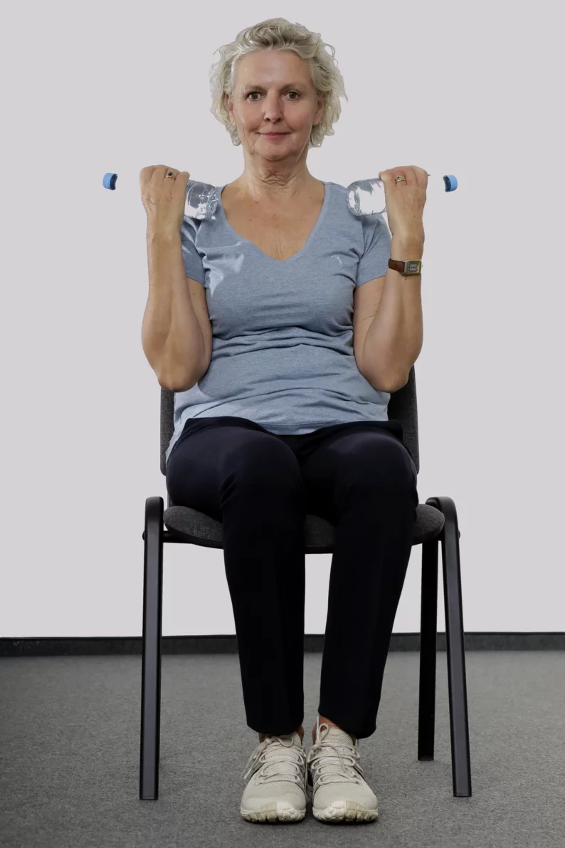 Frau sitzt auf einem Stuhl mit Wasserflaschen in der Hand und zieht die Hände mit den Wasserflaschen an die Schulter