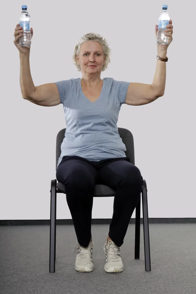 Frau sitzt auf einem Stuhl, hat in jeder Hand eine kleine Wasserflasche und öffnet die angewinkelten Armen zu beiden Seiten